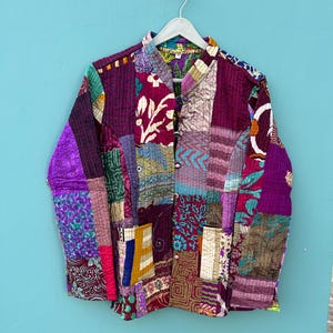 Puede incluir: Una chaqueta acolchada de patchwork multicolor con cuello mandarín y cierre de botones. La chaqueta está hecha de varias telas con patrones florales y geométricos en tonos de morado, rosa, azul, verde y marrón.