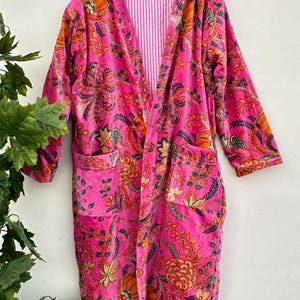 Puede incluir: Una bata rosa vibrante con un estampado floral en naranja, amarillo y verde. La bata tiene un forro a rayas rosa y blanco y dos bolsillos delanteros. La bata está colgada de una percha de madera.