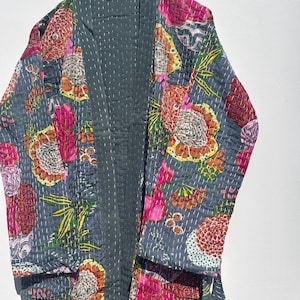 May include: A gray quilted robe with a vibrant floral pattern in shades of pink, orange, and yellow. The robe is hanging on a wooden hanger, showcasing its intricate design and stitching details. The robe's interior is a solid gray color.