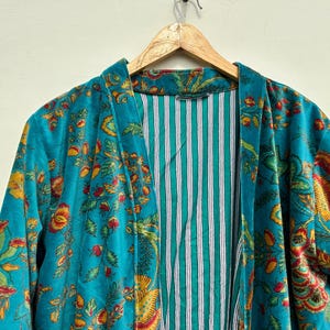 May include: A teal-colored robe with a floral pattern in yellow, red, and green. The robe's interior features a striped pattern in teal and white. The robe is hanging on a wooden hanger.