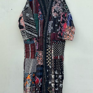 Könnte beinhalten: Ein langer Patchwork-Bademantel mit einer Vielzahl von Mustern und Farben, darunter Blumen-, geometrische und abstrakte Designs. Der Bademantel ist überwiegend schwarz mit Akzenten in Rot, Grün und Weiß. Er hängt an einem Holzbügel.