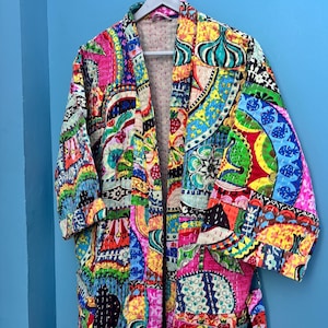 Op de afbeelding: Een levendige patchwork-badjas met een kleurrijk ontwerp. De badjas heeft een mix van roze, gele, groene en blauwe patronen. Hij hangt aan een witte hanger tegen een blauwe muur. De badjas lijkt gewatteerd te zijn.