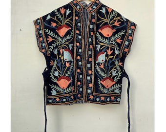 May include: A navy blue, sleeveless embroidered jacket with a mandarin collar. The jacket features a floral design in shades of coral, blue, and gold. It has tie closures and is hanging on a wooden hanger.