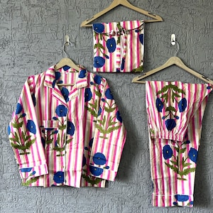 以下が含まれることがあります： ピンクと白のストライプのパジャマセット、青い花の刺繍入り。 セットには、ボタンダウンシャツ、パンツ、お揃いの巾着袋が含まれています。 パジャマは、木製のハンガーにかけられ、質感のある灰色の壁に掛けられています。