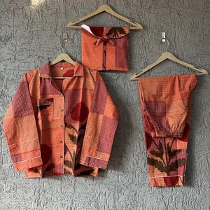 Peut inclure: Ensemble de pyjama trois pièces aux tons orange et rouge, avec un imprimé floral. L'ensemble comprend une chemise boutonnée, un pantalon et une pochette assortie, le tout suspendu à des cintres en bois sur un mur gris texturé.
