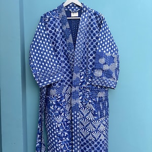 May include: A blue and white patterned robe hanging on a white hanger against a light blue wall. The robe features a variety of patterns, including polka dots, swirls, and floral designs. The robe has a matching belt.