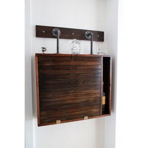 May include: A dark wood cabinet with a fold-down front, mounted to a wall with industrial pipe hardware. The cabinet has a small open shelf on the right side.
