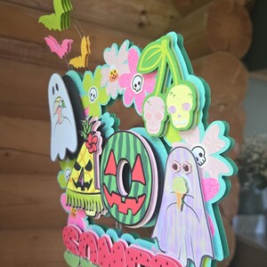 May include: A layered, colorful wreath with a summer theme. It features a watermelon slice, ghosts, skulls, flowers, and the word "Summer" in red. The wreath is held by a thin wire.
