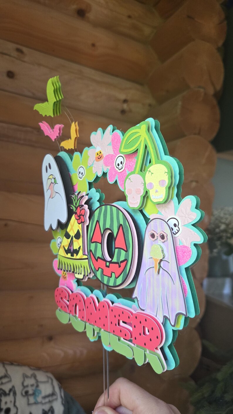 May include: A layered, colorful wreath with a summer theme. It features a watermelon slice, ghosts, skulls, flowers, and the word "Summer" in red. The wreath is held by a thin wire.
