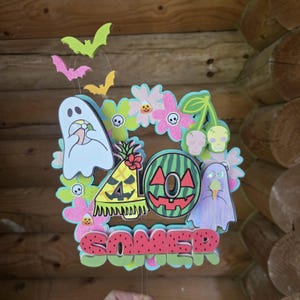 May include: A colorful Halloween-themed decoration featuring a watermelon with a jack-o'-lantern face, ghosts, flowers, bats, and the word "SOMER" in a watermelon-style font. The decoration is made of wood and has a whimsical design.