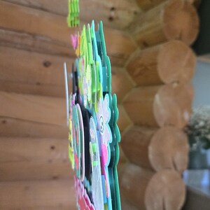 May include: A colorful, layered paper decoration with a floral and cartoon character design. The decoration features various colors, including pink, green, blue, and white. The background is a wooden log cabin wall.