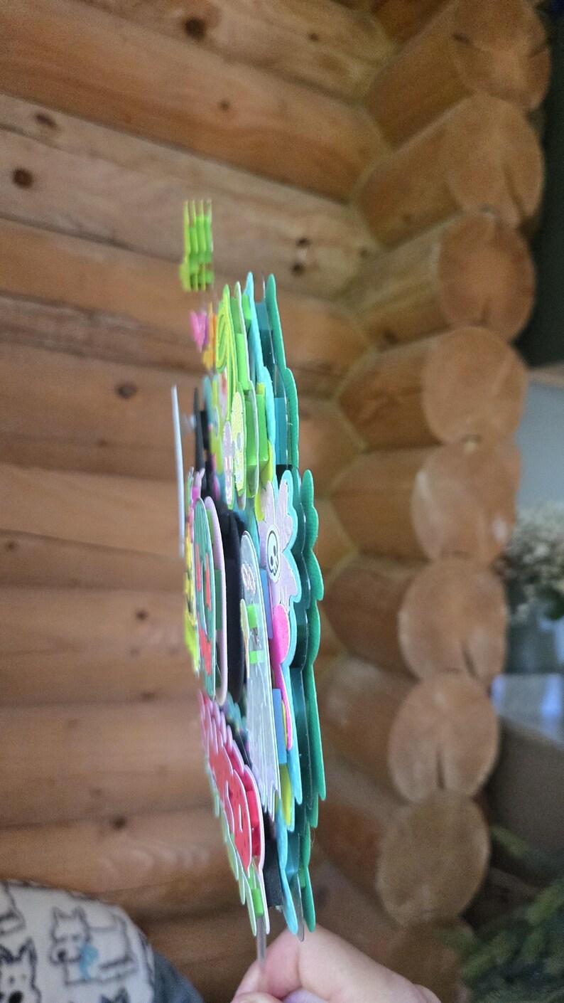 May include: A colorful, layered paper decoration with a floral and cartoon character design. The decoration features various colors, including pink, green, blue, and white. The background is a wooden log cabin wall.