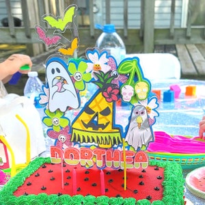 May include: A festive birthday cake with red frosting, green frosting trim, and black seed decorations. The cake is topped with a colorful, Halloween-themed decoration featuring ghosts, bats, flowers, and the name "Dorthea."