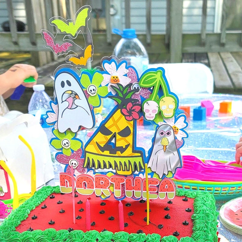 May include: A festive birthday cake with red frosting, green frosting trim, and black seed decorations. The cake is topped with a colorful, Halloween-themed decoration featuring ghosts, bats, flowers, and the name "Dorthea."
