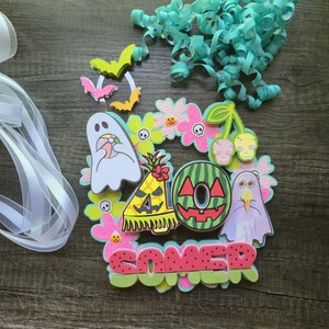 May include: A colorful Halloween-themed wreath with a variety of cut-out shapes. The wreath includes ghosts, bats, flowers, and the word "SOMER" in red letters. Teal ribbon and curly paper are also present.