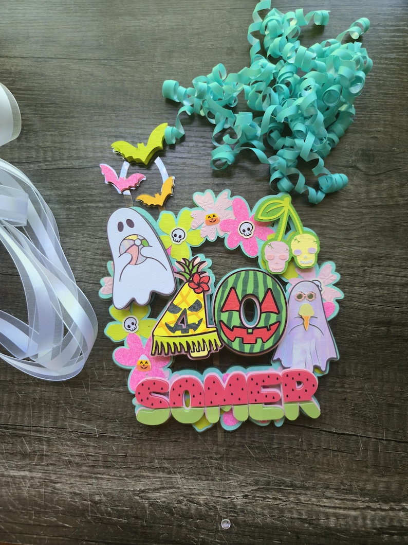 May include: A colorful Halloween-themed wreath with a variety of cut-out shapes. The wreath includes ghosts, bats, flowers, and the word "SOMER" in red letters. Teal ribbon and curly paper are also present.