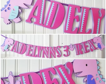Può includere: Striscione a tema dinosauro rosa e viola con il nome "Adelynns 3 Rex" in lettere rosa con un motivo blu e bianco. Lo striscione è infilato su un nastro viola e presenta ritagli di dinosauri.