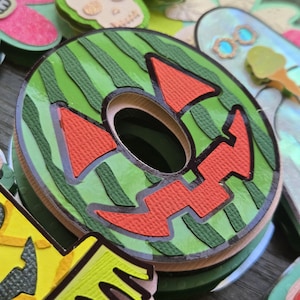 May include: A layered, circular Halloween decoration with a watermelon design. The green outer ring has vertical stripes, and the center features a jack-o'-lantern face in red and black. Other decorative elements are visible in the background.
