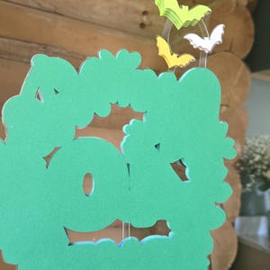 May include: A green, cloud-shaped decoration with cut-out details, held by clear strings. Above it are three bat-shaped decorations in yellow, white, and green. The background is a wooden wall.
