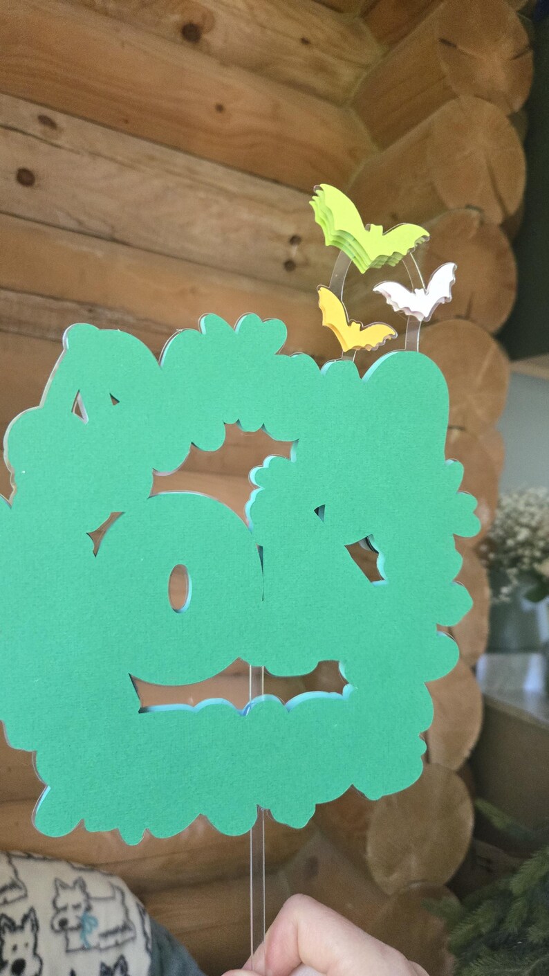 May include: A green, cloud-shaped decoration with cut-out details, held by clear strings. Above it are three bat-shaped decorations in yellow, white, and green. The background is a wooden wall.