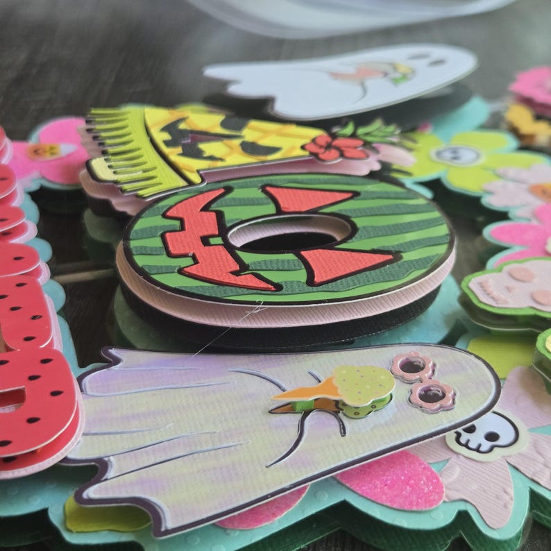 May include: Close-up of colorful layered paper cutouts, including a green and red watermelon-themed letter "O", a yellow pineapple, and ghost-shaped figures with ice cream cones. The cutouts are arranged in a decorative, festive style.