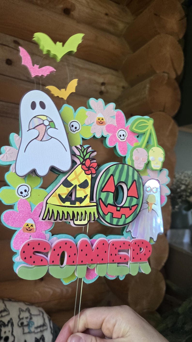 May include: A colorful Halloween-themed decoration with the word "SOMER" in watermelon-style letters. The design includes ghosts, bats, flowers, and a pineapple with the number "40".