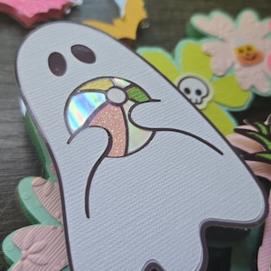May include: A light blue ghost-shaped cutout with a colorful beach ball design. The ghost is surrounded by decorative flowers, bats, and other Halloween-themed cutouts. The image has a shallow depth of field.