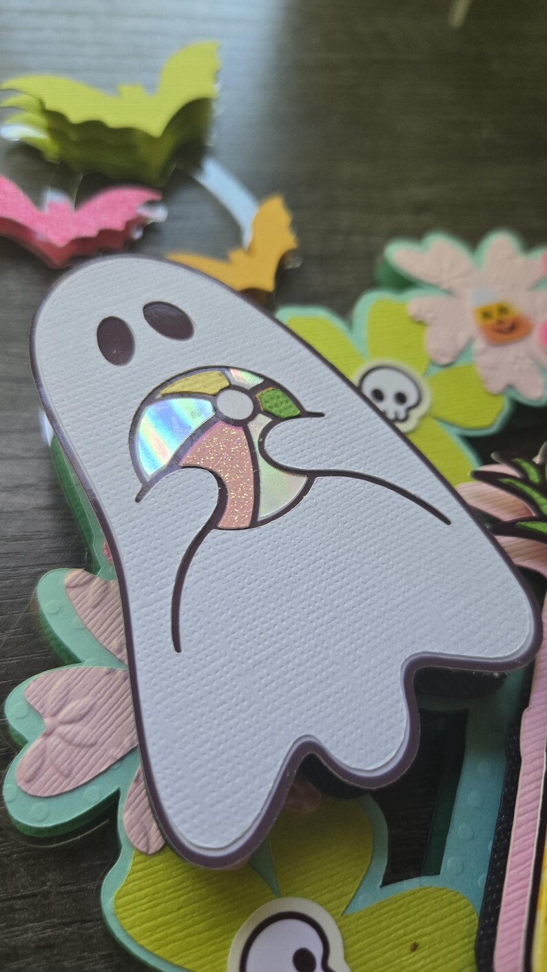 May include: A light blue ghost-shaped cutout with a colorful beach ball design. The ghost is surrounded by decorative flowers, bats, and other Halloween-themed cutouts. The image has a shallow depth of field.