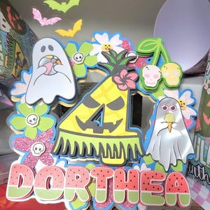 May include: A layered paper decoration with a watermelon-themed name, "Dorthea." The design includes a pineapple with the number 4, ghosts, flowers, and bats. The color palette features pink, green, yellow, and blue accents, creating a festive, celebratory theme.