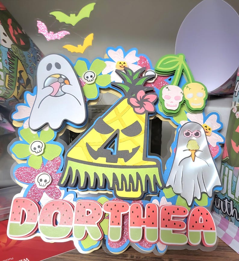 May include: A layered paper decoration with a watermelon-themed name, "Dorthea." The design includes a pineapple with the number 4, ghosts, flowers, and bats. The color palette features pink, green, yellow, and blue accents, creating a festive, celebratory theme.
