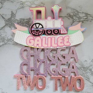 May include: A pink and white train cake topper with the name "GALILEE" and the words "CHUGGA CHUGGA TWO TWO" in pink glitter letters.