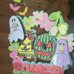 May include: A colorful, layered cake topper with a summer and Halloween theme. The design includes ghosts, bats, flowers, a watermelon, and the word "SOMER". The topper is held by a clear stick.