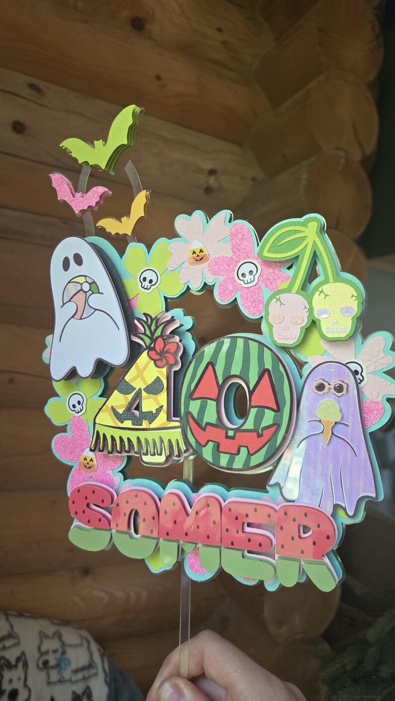 May include: A colorful, layered cake topper with a summer and Halloween theme. The design includes ghosts, bats, flowers, a watermelon, and the word "SOMER". The topper is held by a clear stick.