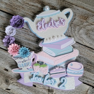 May include: A layered paper craft featuring a teapot, books, teacup, strawberries, and a cake. The design includes the text "Electra's" and "Tea for 2". The color palette includes white, light blue, pink, purple, and green.