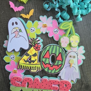 May include: A colorful, layered decoration with a summer and Halloween theme. It features a watermelon-themed letter O, ghosts, flowers, bats, and the word "SOMER" in watermelon colors. Decorative ribbons are in the background.