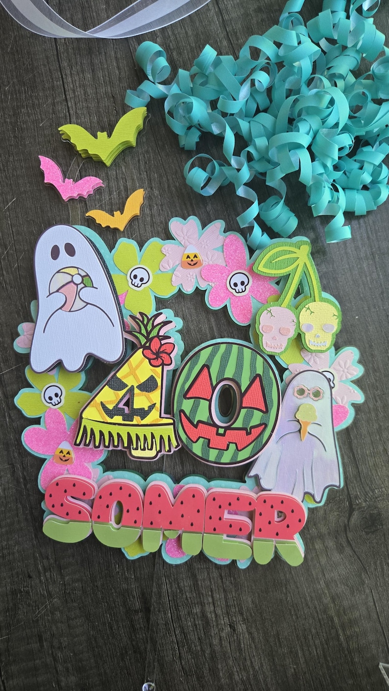May include: A colorful, layered decoration with a summer and Halloween theme. It features a watermelon-themed letter O, ghosts, flowers, bats, and the word "SOMER" in watermelon colors. Decorative ribbons are in the background.
