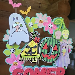 May include: A colorful, layered decoration with a summer theme. It features a watermelon slice, ghosts, flowers, and the word "SOMER" in red letters. Bats and other decorative elements are also included.