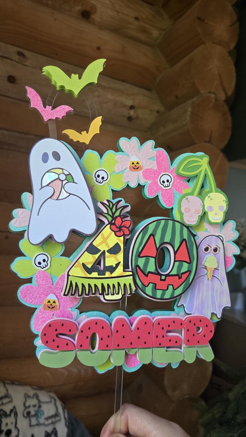 May include: A colorful, layered decoration with a summer theme. It features a watermelon slice, ghosts, flowers, and the word "SOMER" in red letters. Bats and other decorative elements are also included.