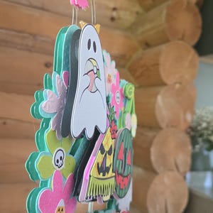 May include: A colorful, layered paper decoration with a ghost, flowers, and a watermelon design. The word "Summer" is displayed in red letters. The decoration has a whimsical, festive appearance, perfect for seasonal decor.