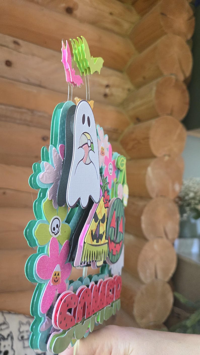 May include: A colorful, layered paper decoration with a ghost, flowers, and a watermelon design. The word "Summer" is displayed in red letters. The decoration has a whimsical, festive appearance, perfect for seasonal decor.