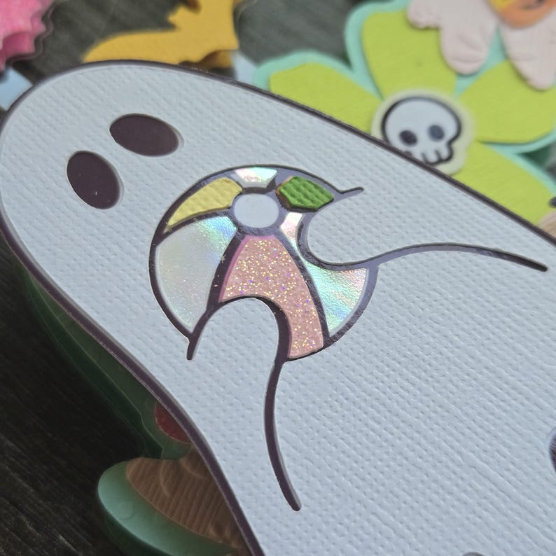 May include: A light blue ghost-shaped paper cutout with two dark purple eye holes. The ghost has a colorful, iridescent umbrella design with pink glitter accents. Other Halloween-themed cutouts are in the background.