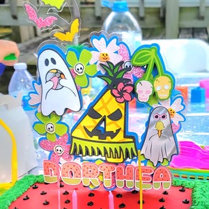 May include: A festive cake with a watermelon design, topped with a colorful Halloween-themed decoration. The decoration includes bats, ghosts, flowers, and a pineapple with the name "ORTHEA". Pink candles are inserted into the cake.