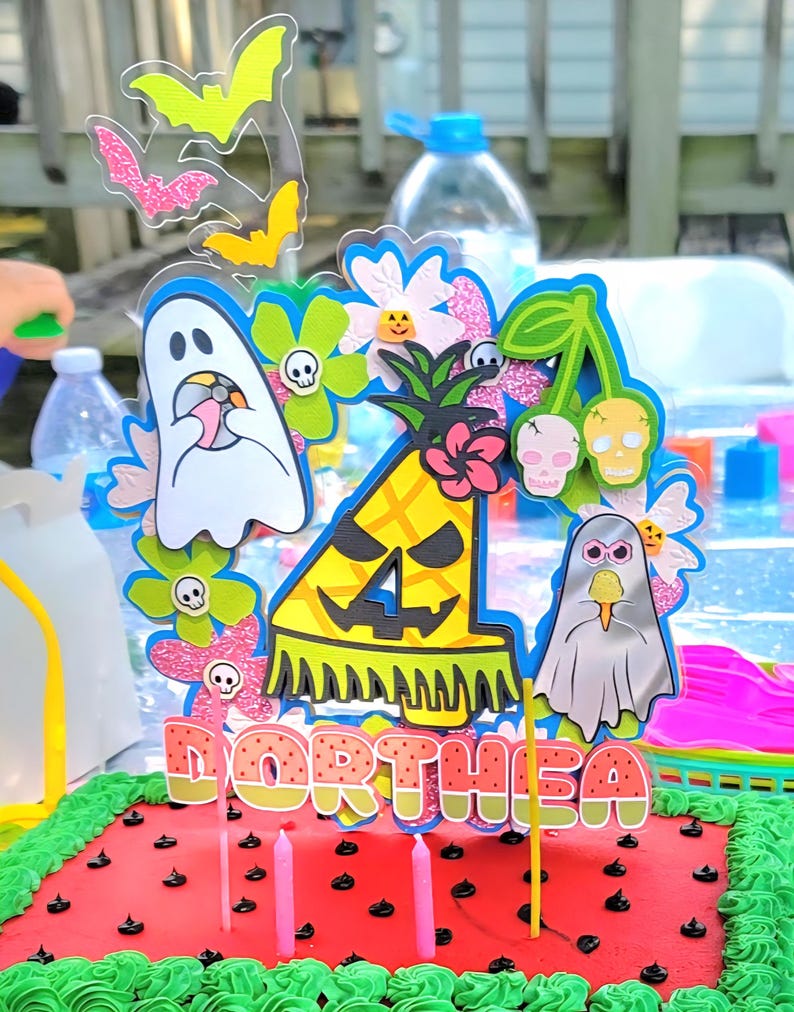 May include: A festive cake with a watermelon design, topped with a colorful Halloween-themed decoration. The decoration includes bats, ghosts, flowers, and a pineapple with the name "ORTHEA". Pink candles are inserted into the cake.