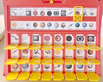 Könnte beinhalten: Ein rotes und gelbes Guess Who?-Spiel mit Weltraumkarten. Das Spielbrett hat gelbe Klappen und Karten mit Bildern von Planeten, Sternen und anderen Himmelskörpern. Der Titel des Spiels ist oben aufgedruckt.