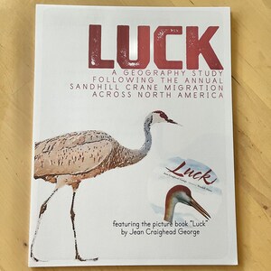 May include: A book cover with a red title "LUCK" and a drawing of a sandhill crane. The text below the title reads "A geography study following the annual sandhill crane migration across North America." The cover also features the text "featuring the picture book "Luck" by Jean Craighead George."