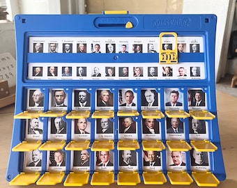 Könnte beinhalten: Ein blaues und gelbes Spielbrett aus Plastik mit dem Titel "Guess Who?" und einer Schiebefläche, um Bilder von US-Präsidenten zu enthüllen. Das Spielbrett hat einen gelben Schieberegler mit den Zahlen 0-4.