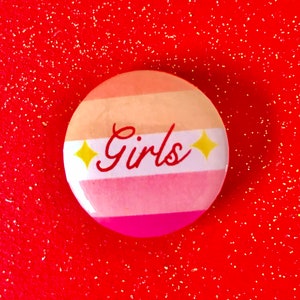 Puede incluir: Un botón redondo con un diseño a rayas rosa, blanco y naranja. La palabra "Girls" está escrita en letras blancas cursivas en el botón. Hay dos estrellas amarillas a cada lado de la palabra.