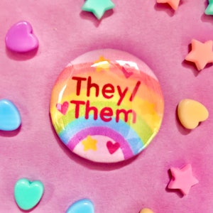 Könnte beinhalten: Ein regenbogenfarbener Knopf mit dem Text "They/Them" in roten Buchstaben und einem Herzdesign. Der Knopf ist von kleinen Herzen und Sternen umgeben.