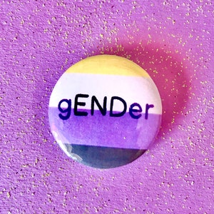 May include: A round button with the word "gender" printed in black on a background of yellow, white, purple, and black stripes.