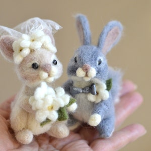 Könnte beinhalten: Zwei Filz-Hochzeitsfiguren in Form von Hasen. Die Braut-Häschen trägt einen weißen Schleier und hält einen Strauß weißer Blumen. Der Bräutigam-Hase trägt eine graue Fliege und hält eine einzelne weiße Blume.
